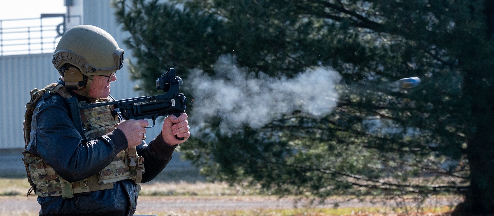 A side profile of a man in full military uniform and protective gear, identified as Maj. Gen. D. Scott Durham, shoots a grenade launcher. A projectile can be seen leaving the weapon, at the far right edge of the photo.