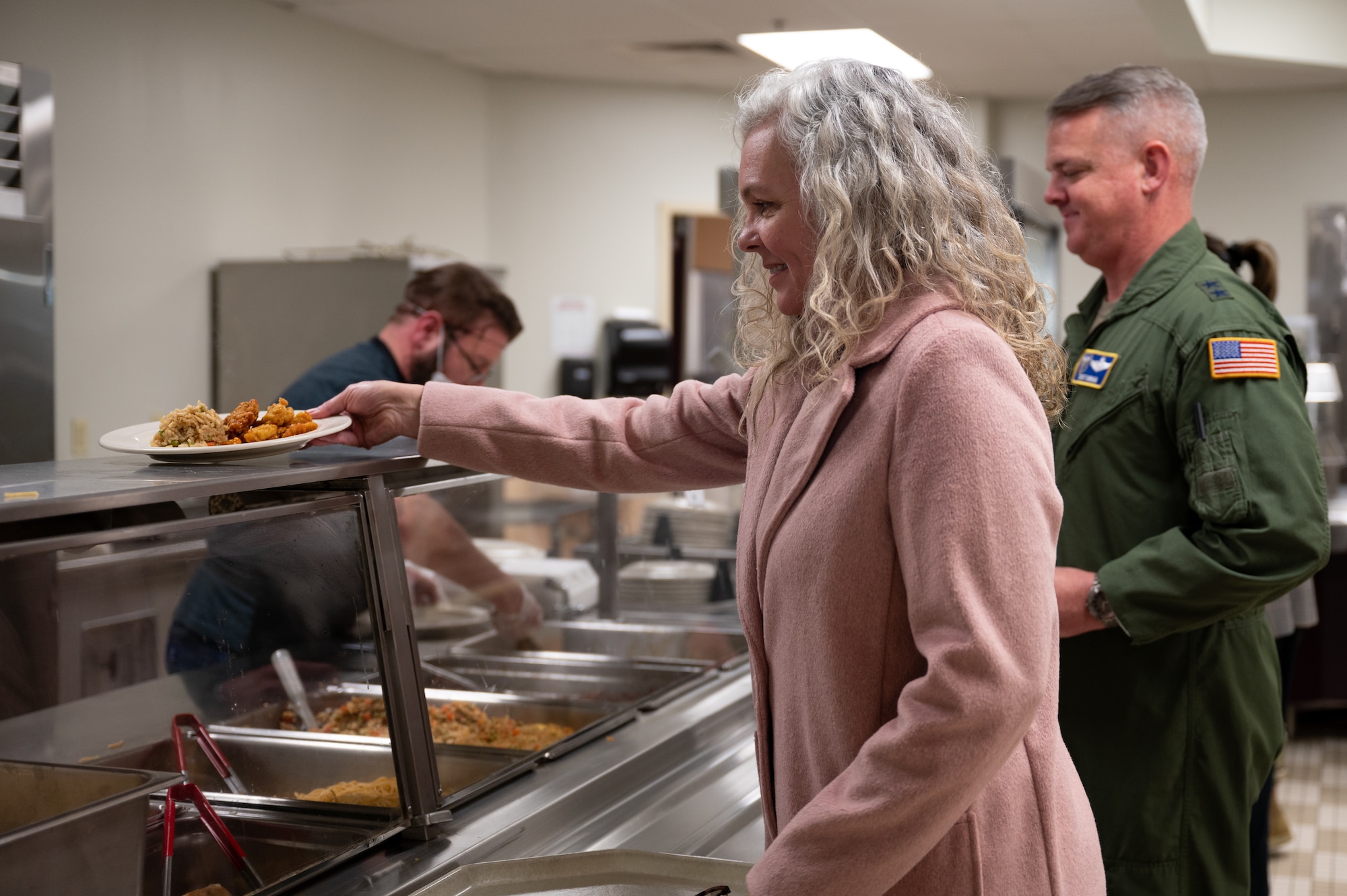 A side profile of Jennifer Durham grabbing a plate of food. She is dressed in a pink coat and has medium-length gray hair. Maj. Gen. Durham is visible in the background, wearing a flight suit.