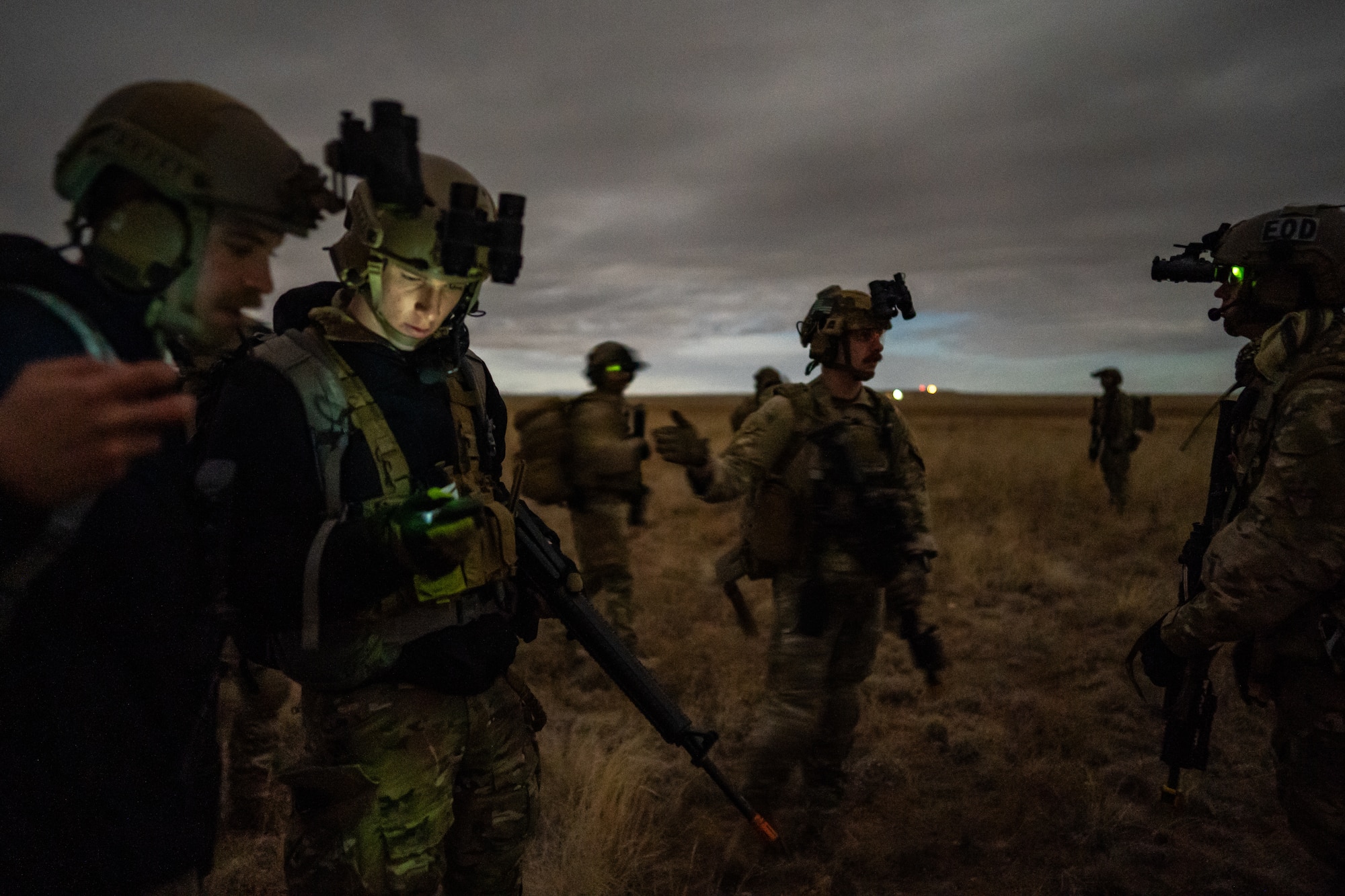 Seven men armed with rifles in military uniform and equipment walk, talk, and look at equipment on an open prairie.