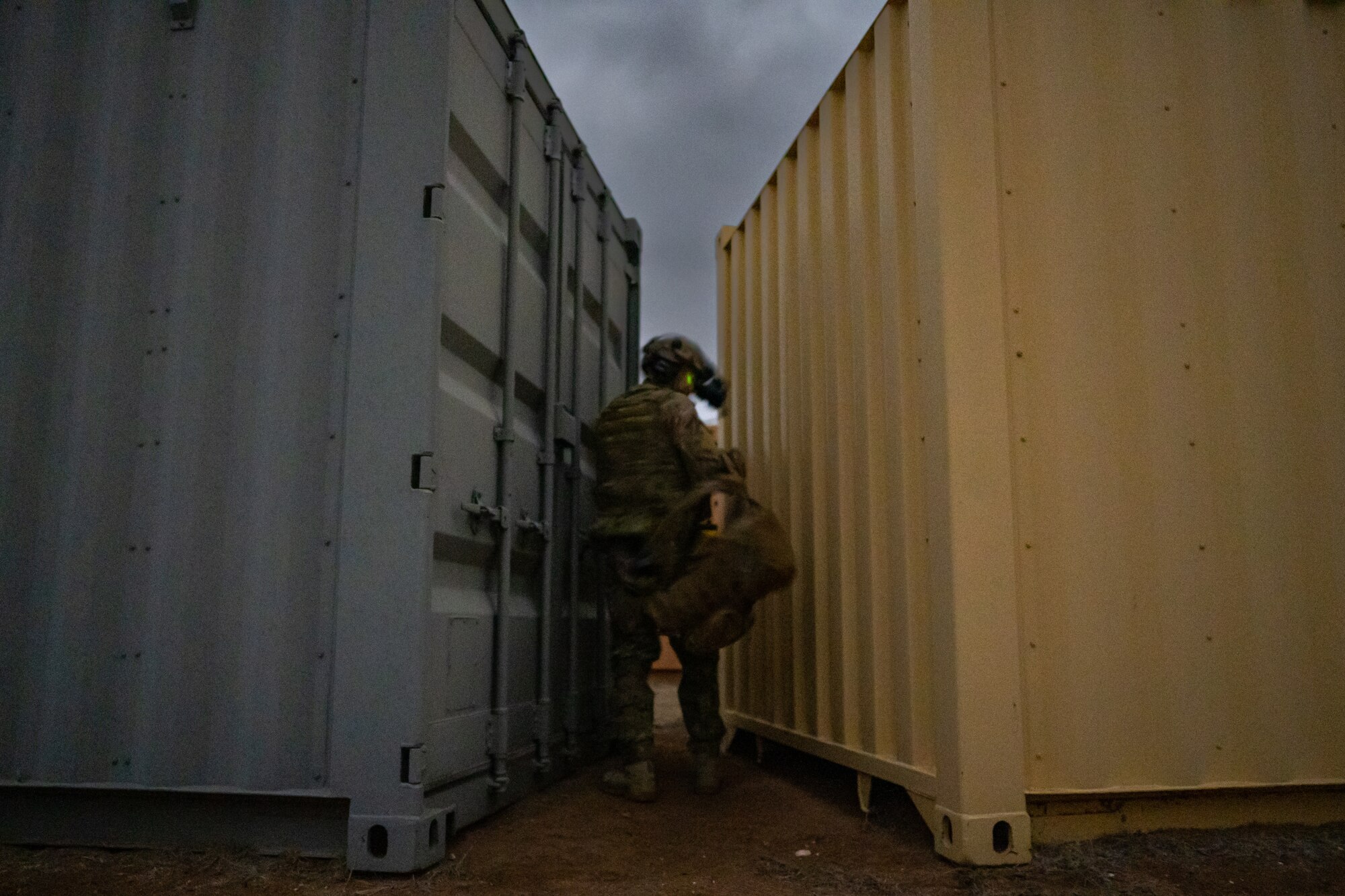 A man in military uniform and equipment removes his backpack in between two shipping containers with a narrow gap between them.