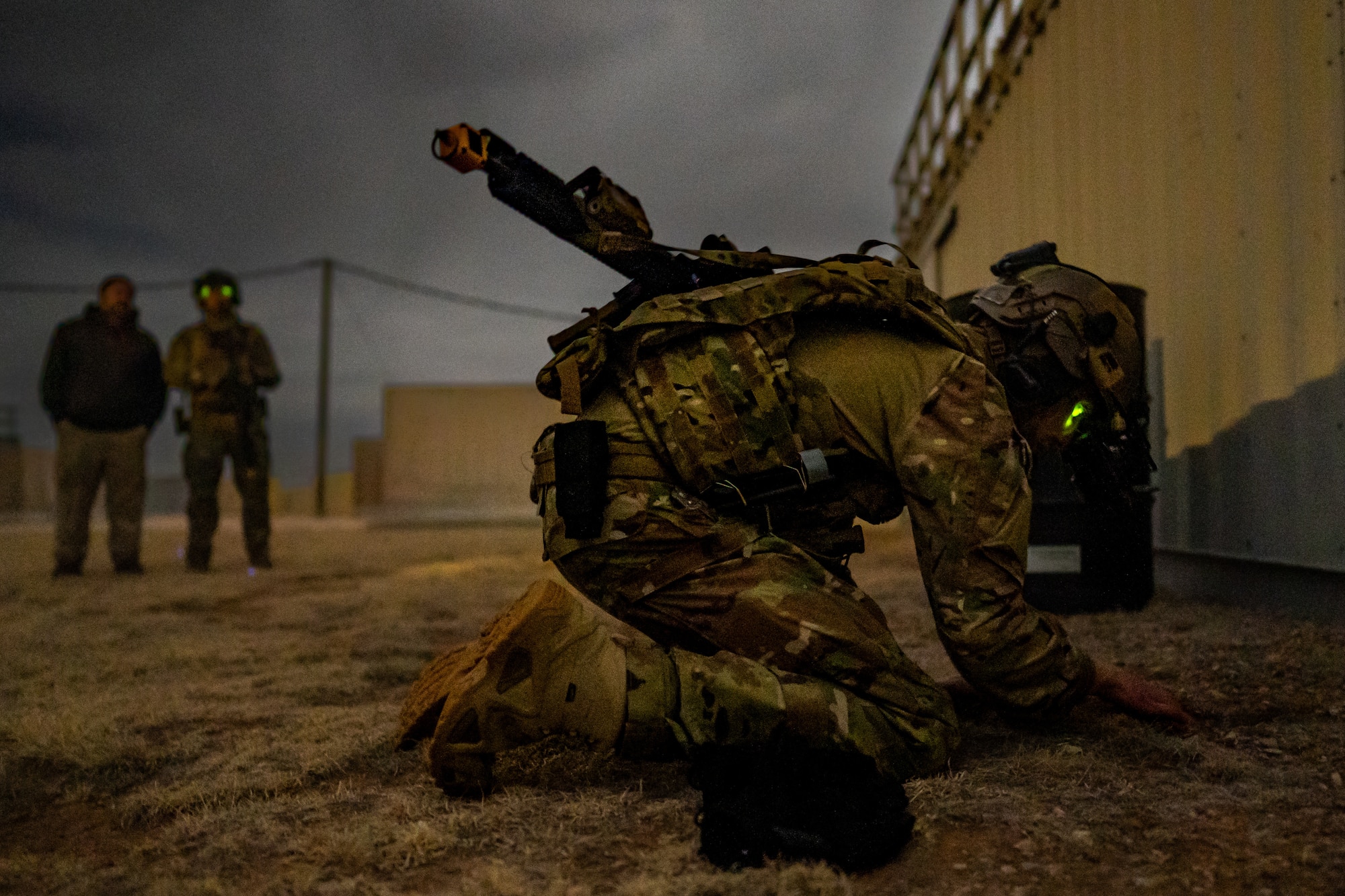 A man in with a rifle in military uniform and equipment kneels on the ground outside a building, digging with his hand. Two men stand in the distance watching him.