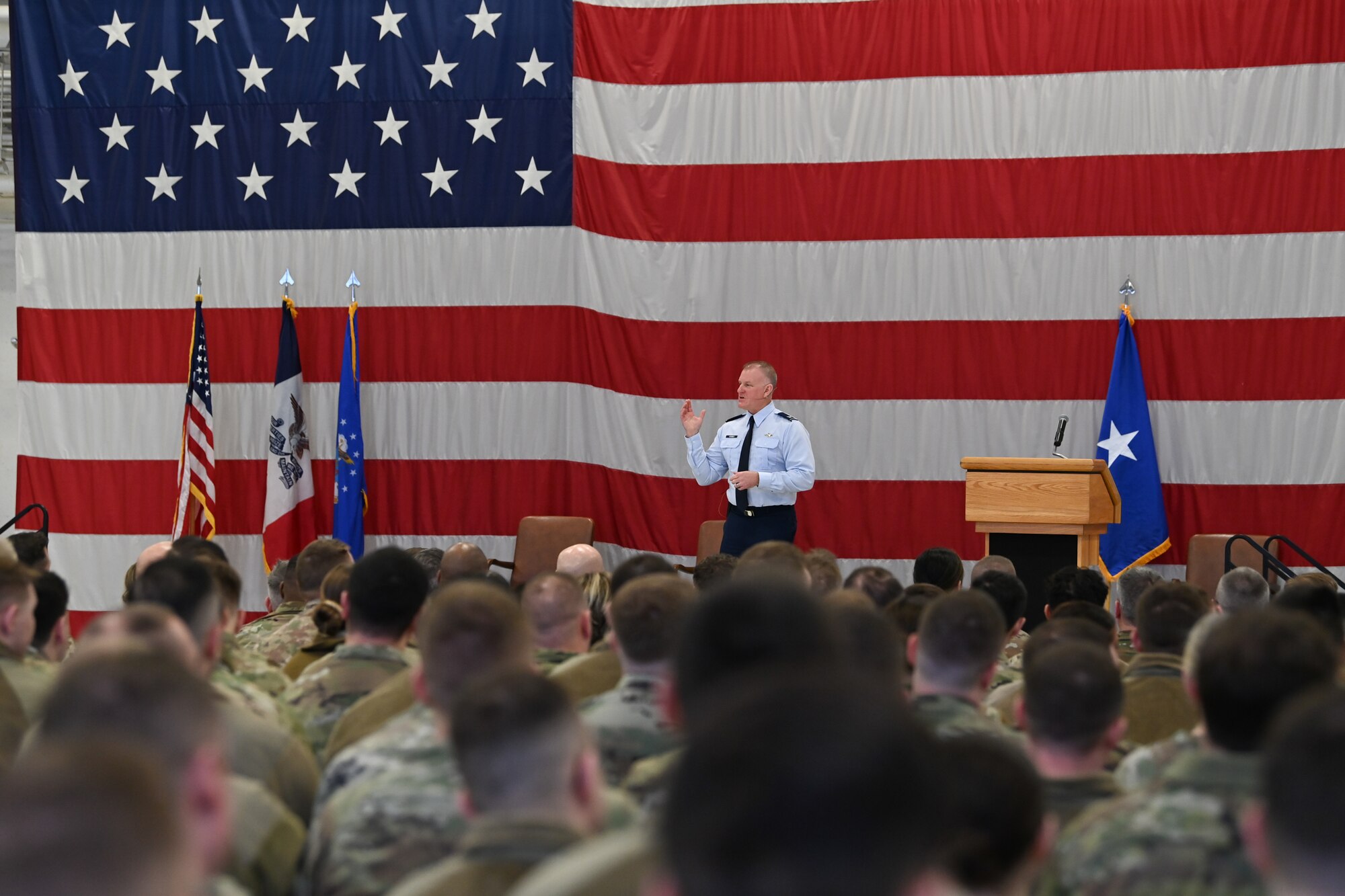 Air Force colonel in blue dress uniform stands in front of large American flag addressing a crowd of Airmen