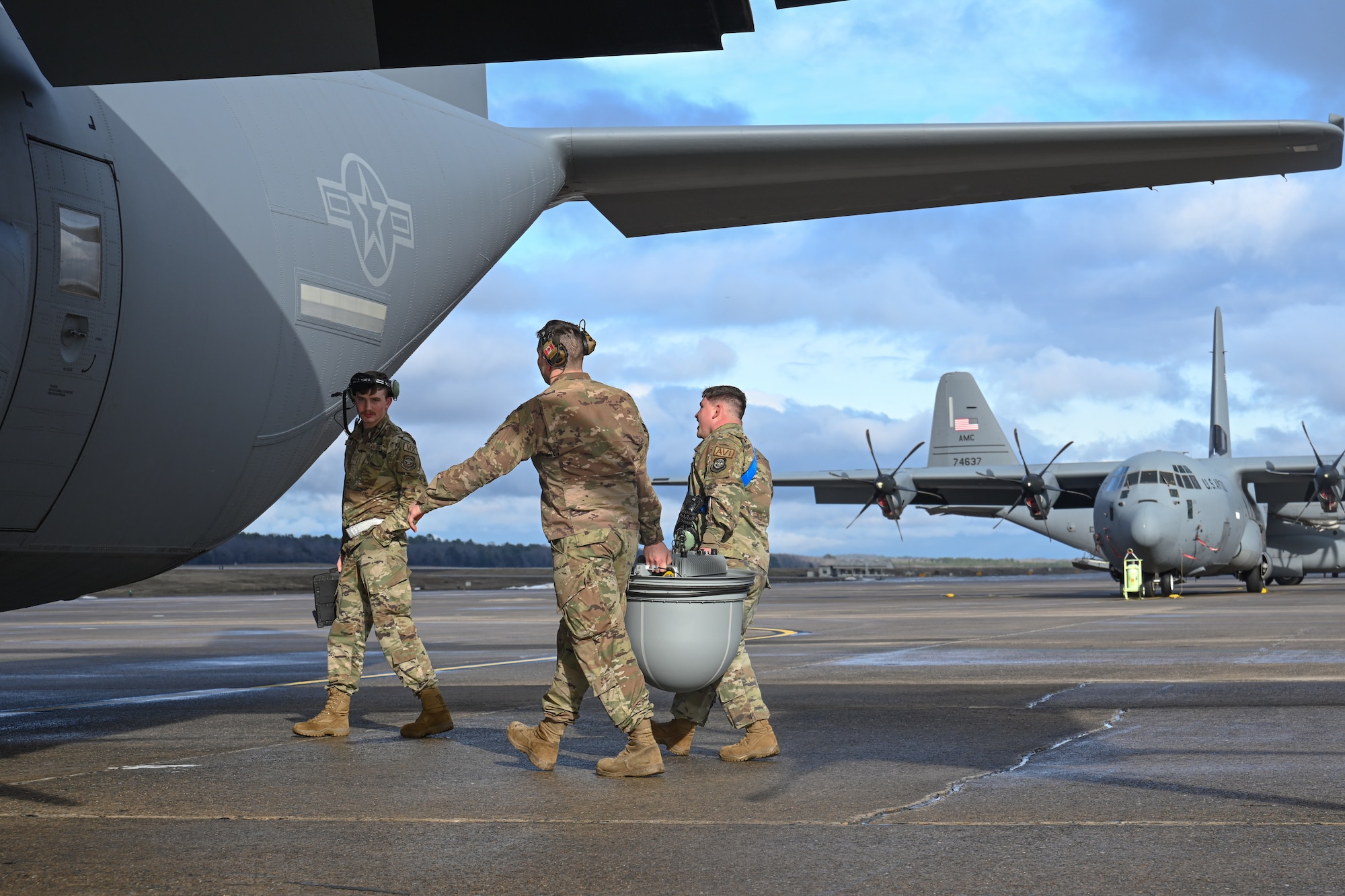 Two Airmen carry a communications pod toward a C-130J as another Airman walks in front of them and watches.