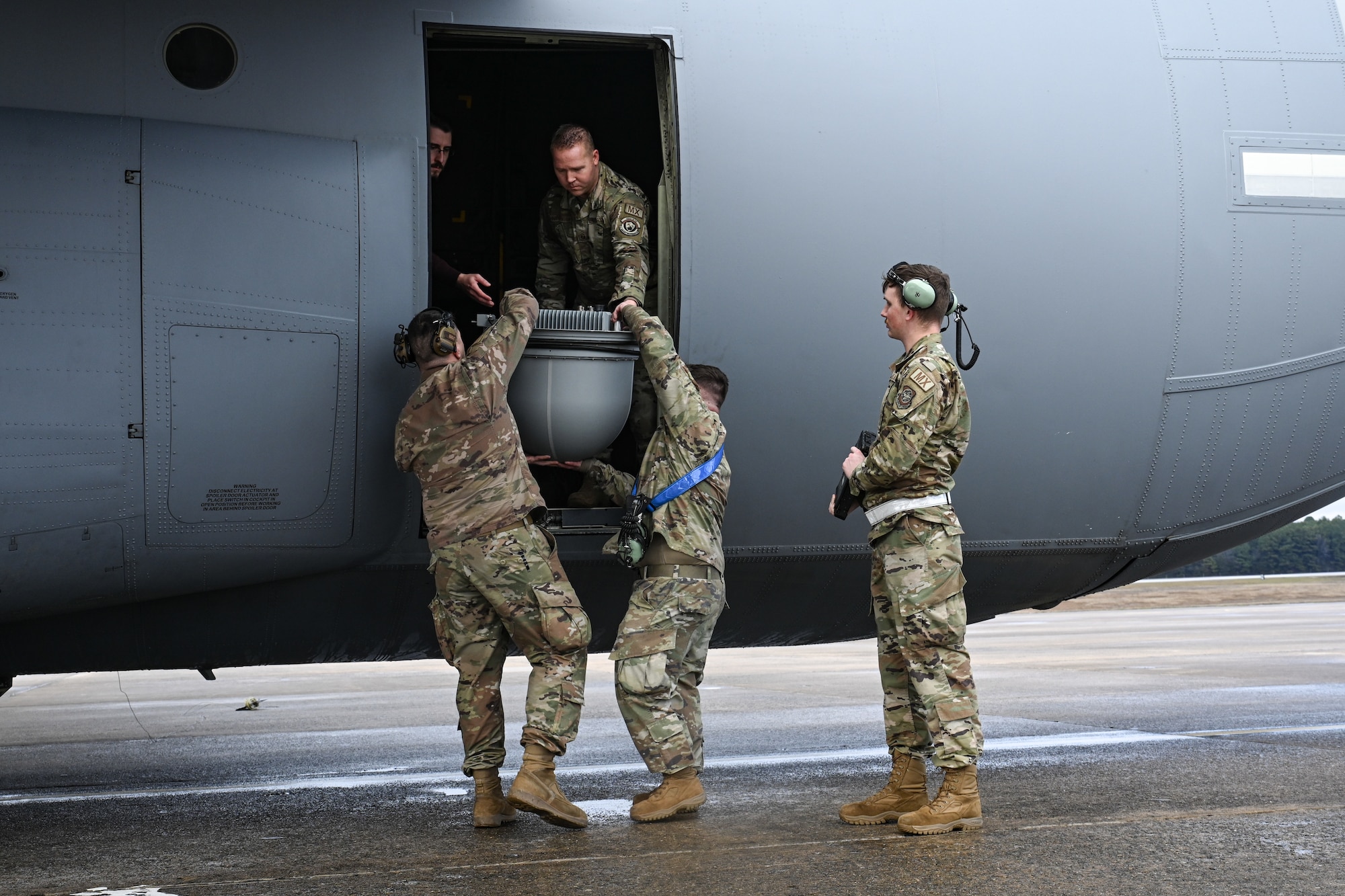 Two Airmen hoist a communications pod up to two Airmen throw the side door of a C-130J side cargo bay door. Another Airman stands to the side and watches over the transfer.