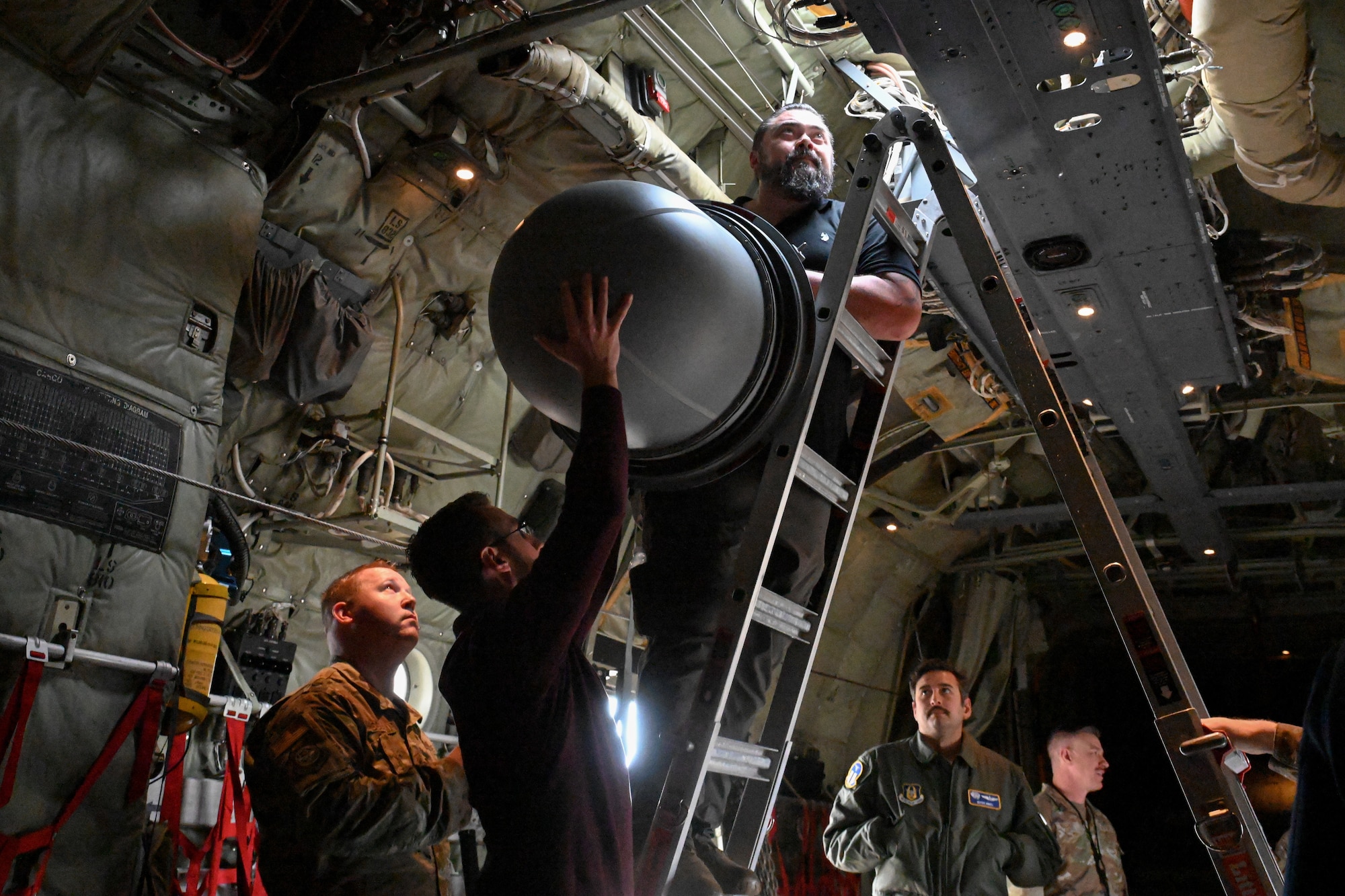 A man hoists a communications pod up to another man standing on a ladder in a C-130J cargo bay. Three Airmen stand around them and watch.