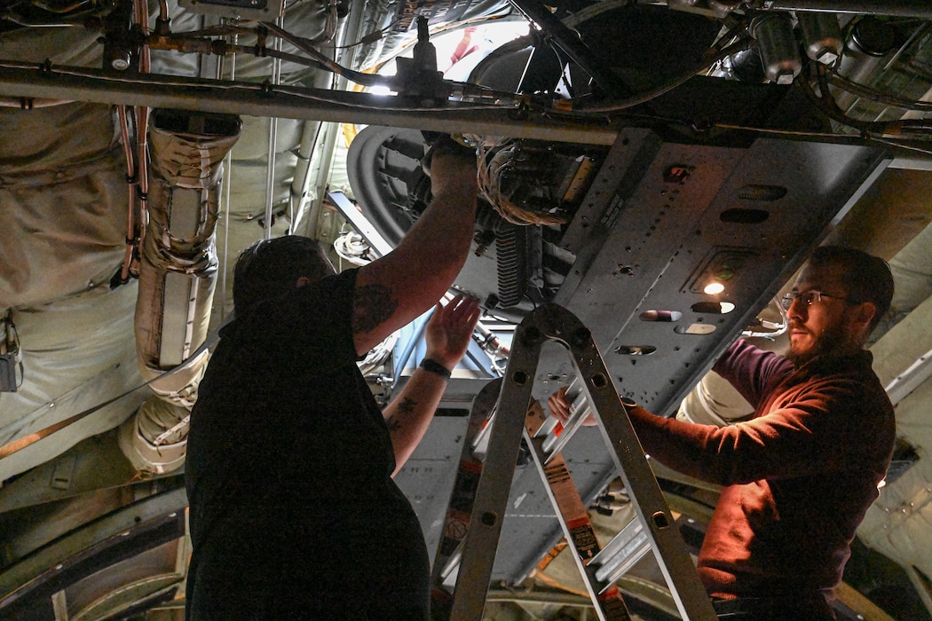 Two men standing on a ladder handle a communication pod in a C-130J aircraft.