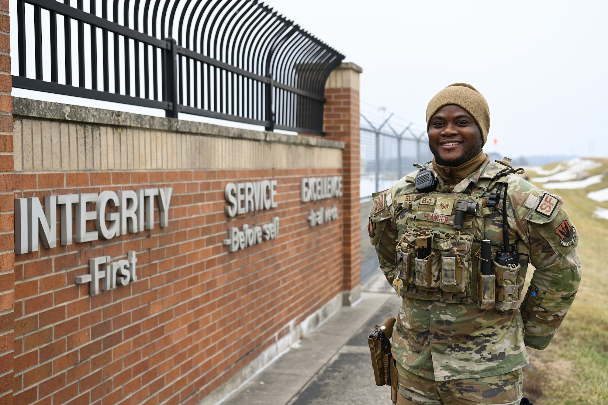 Airman in battle uniform poses for photo outside by a wall bearing the core values of United States Air Force
