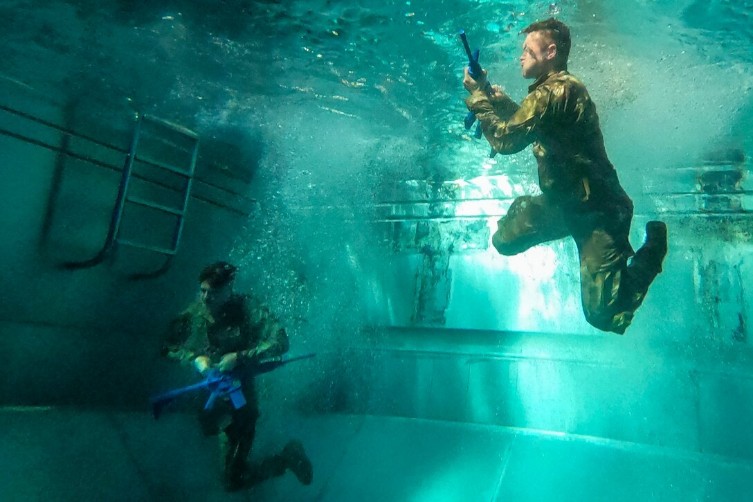 Soldiers swim next to each other underwater in the pool carrying weapons.