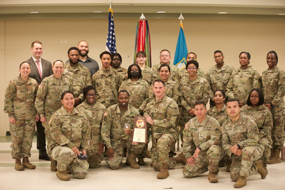 A group of soldiers smile and pose for a photograph. The soldier in the center holds a plaque.