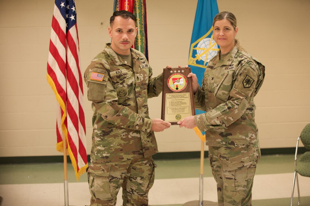 A soldier stands and receives a plaque from another uniformed soldier.