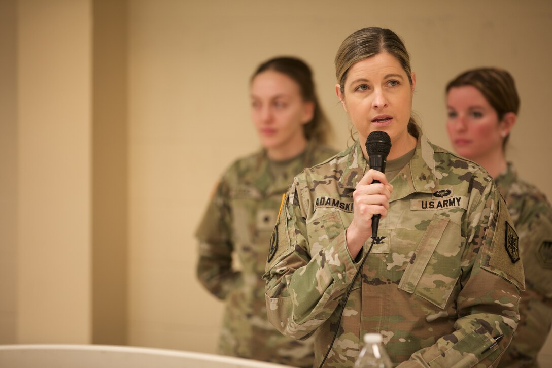 A uniformed soldier addresses a group using a handheld microphone.