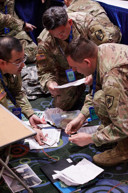 Three soldiers sort a variety of documents and computer peripherals while kneeling on the ground.