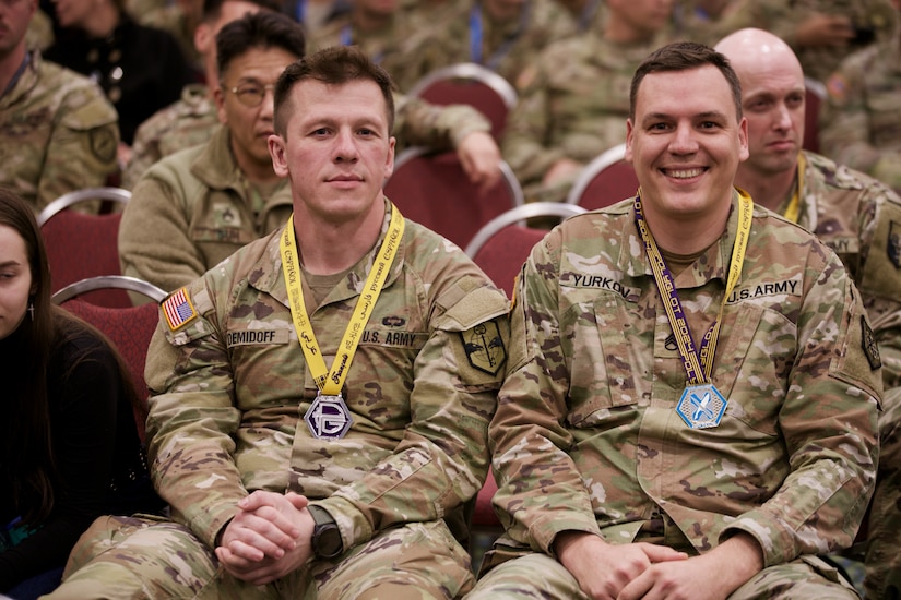 Two soldiers sit next to each other in a crowded room. They have yellow ribbons with medals around their necks.