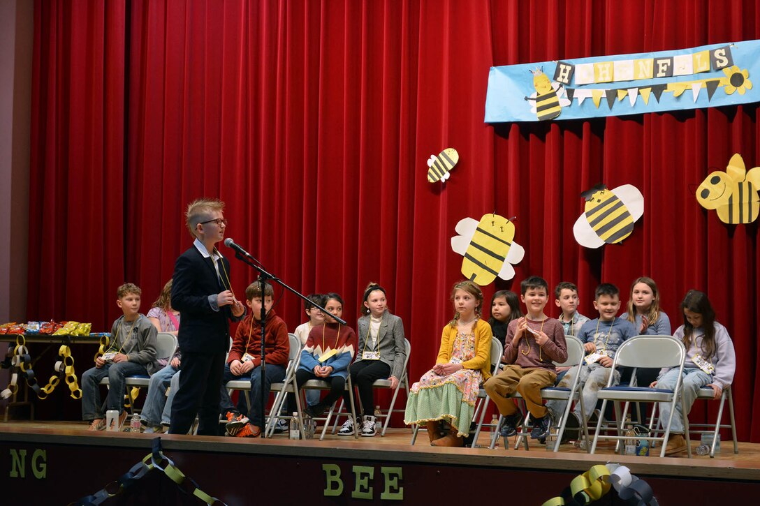 A standing child speaks into a microphone on a stage where other kids sit. Cutout bumblebees adorn a red backdrop curtain.