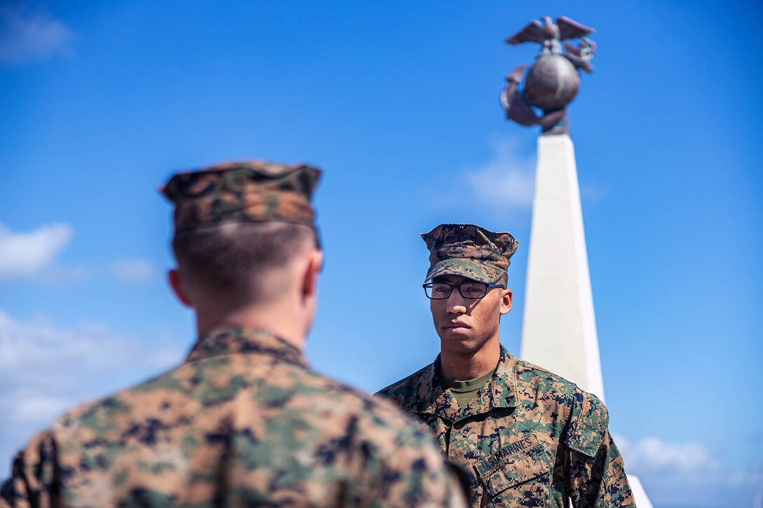Two Marines face each other outdoors in front of a sculpture of the eagle, globe and anchor.