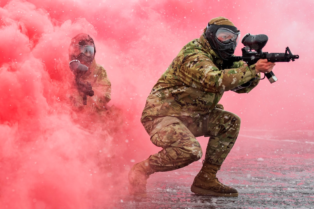 An Air National Guardsman emerges from a thick plume of red smoke wearing a protective mask and holding a weapon, with a soldier behind him.