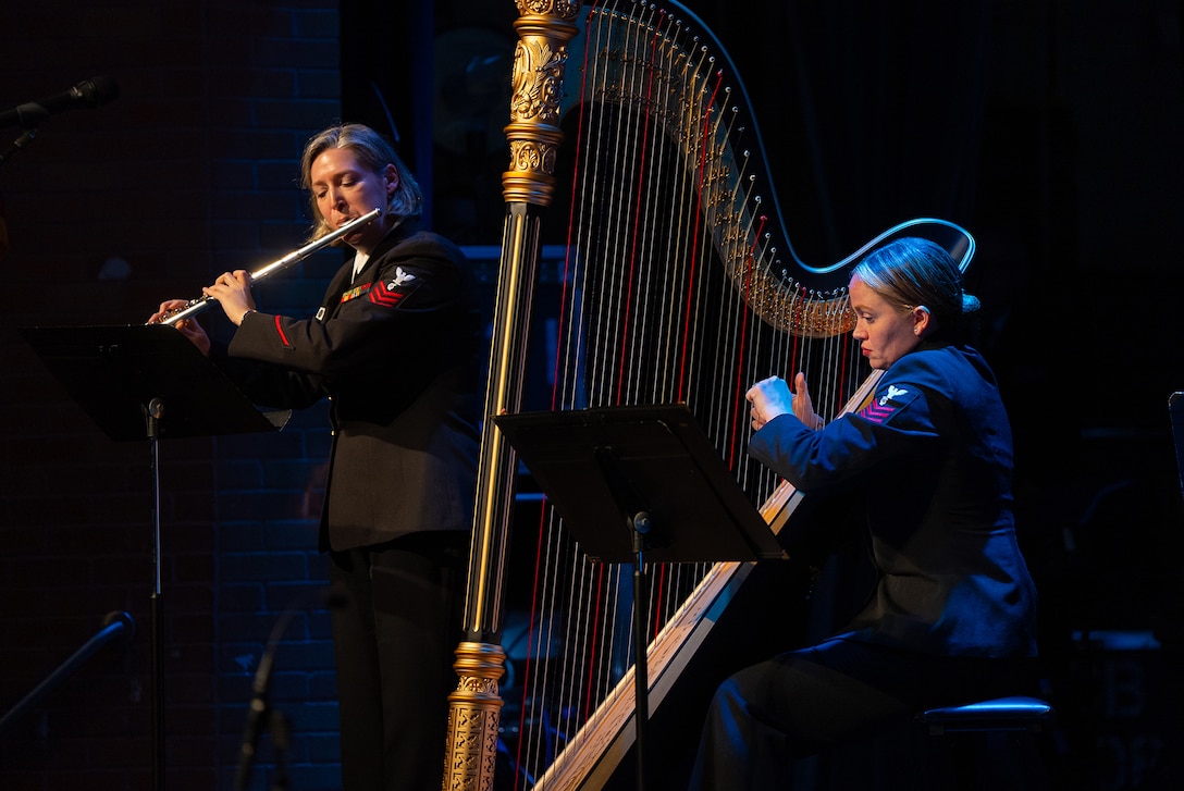 A uniformed sailor plays a flute while a second plays a harp during a performance.