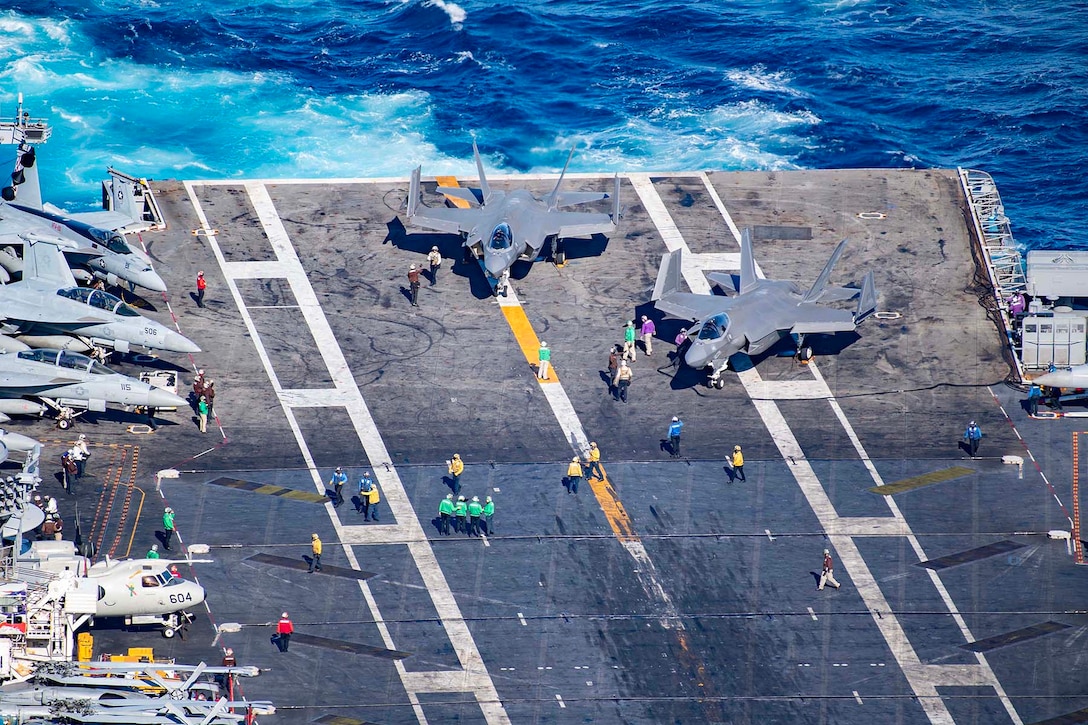 An overhead view of two military aircraft preparing to launch from the flight deck of a Navy ship in open water during daylight.