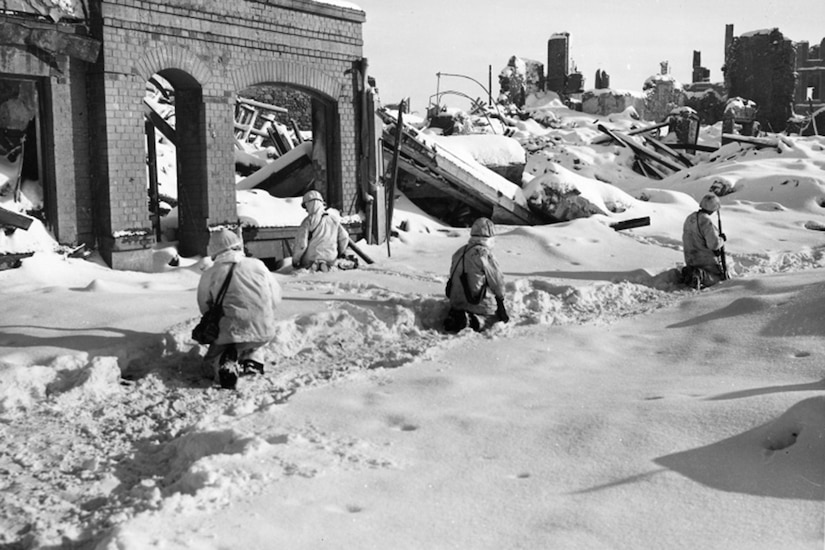 Service members kneel in snow beside destroyed buildings.