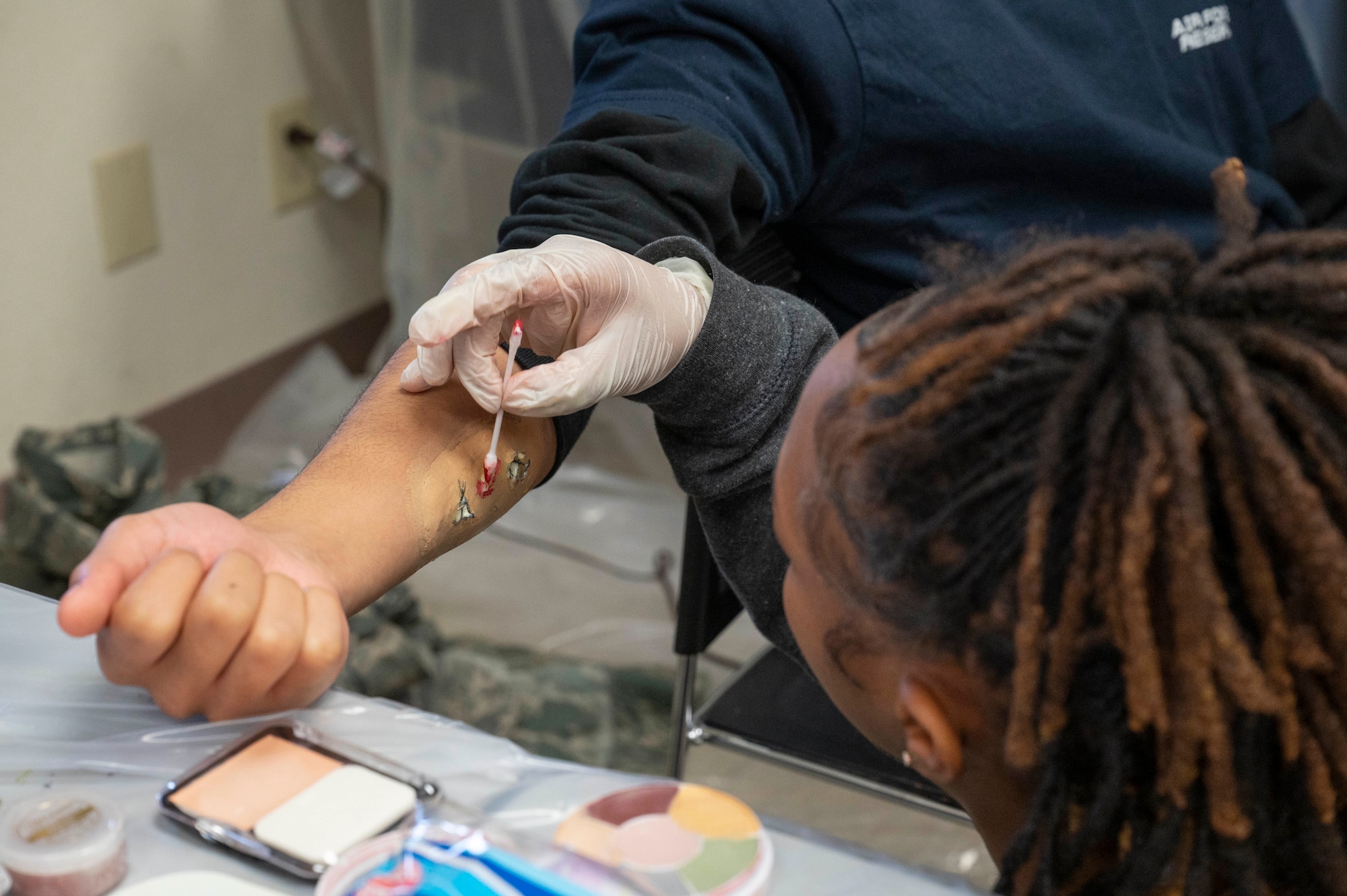 Injury-like makeup is applied onto a trainee's right forearm, resembling a bullet wound.