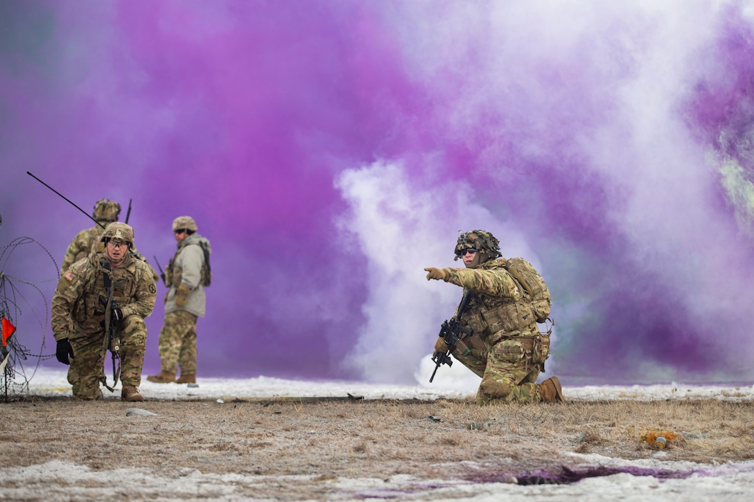 Two soldiers kneel while two soldiers talk behind them as clouds of purple smoke fill the background.