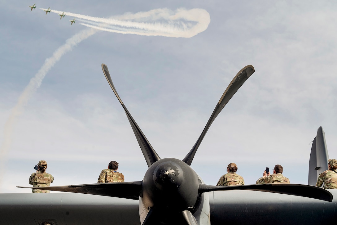 A propeller is pictured in the foreground as airmen sit on the back of the aircraft taking pictures of four aircraft performing a demonstration overhead.