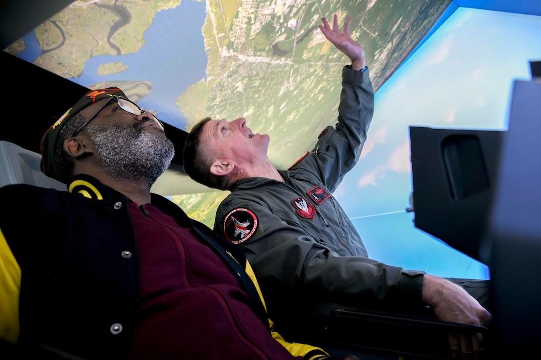 An airman points to a virtual map while sitting at an angle next to a veteran in a flight simulator.