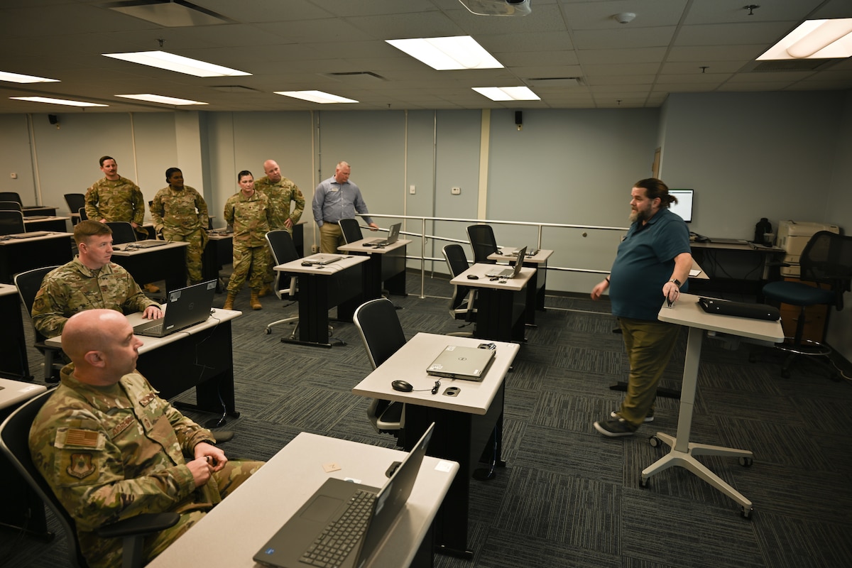 Two Airmen sit in chairs and listen to speaker.
