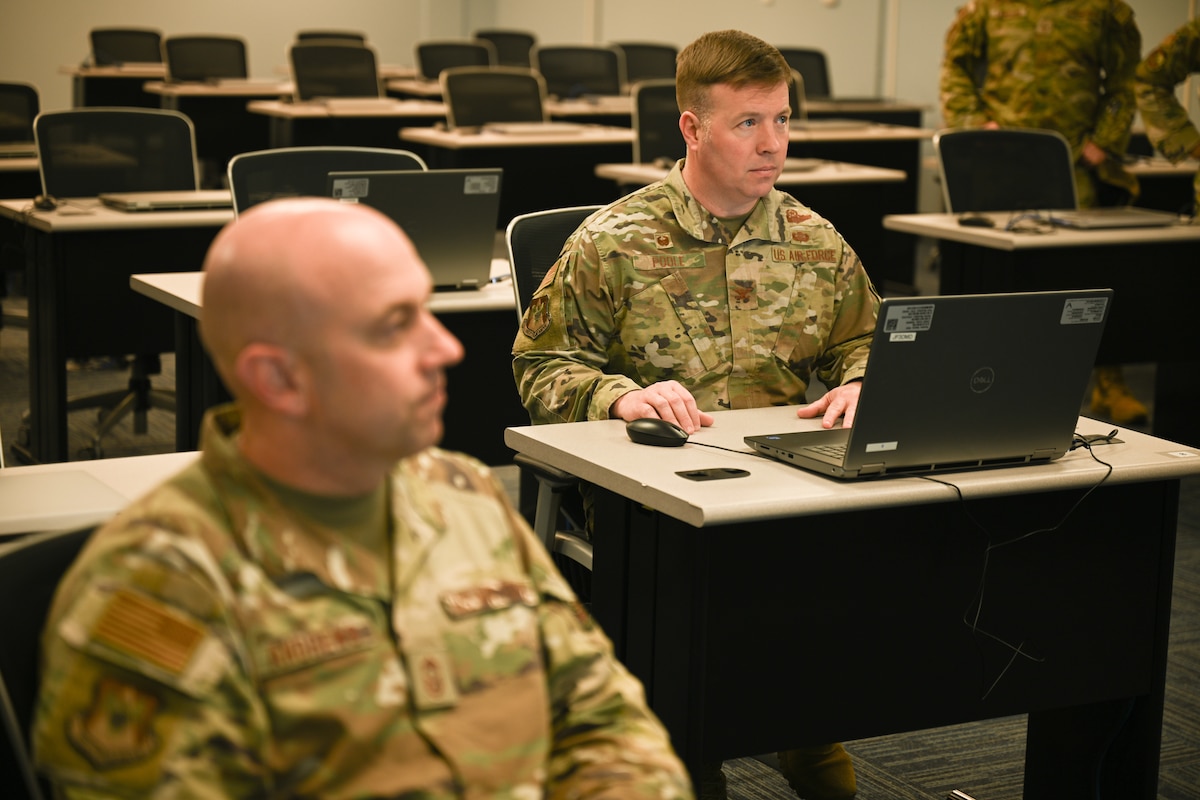 Two Airmen sit in chairs and listen to speaker.