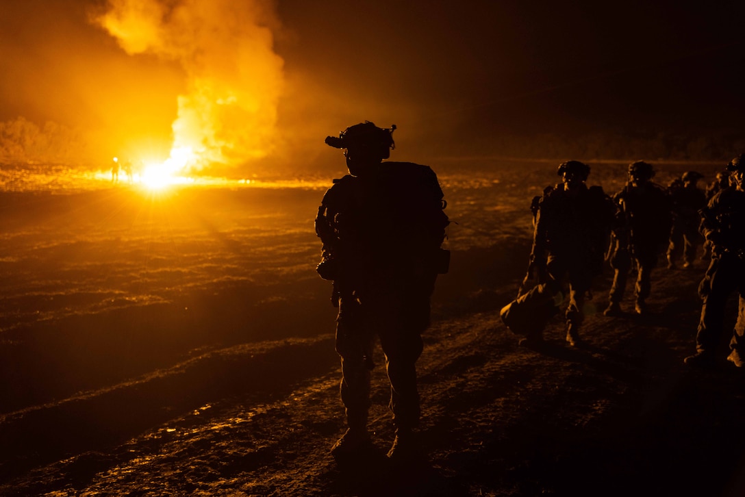 Marines walk along a dirt road at night as a large fire burns in the background.