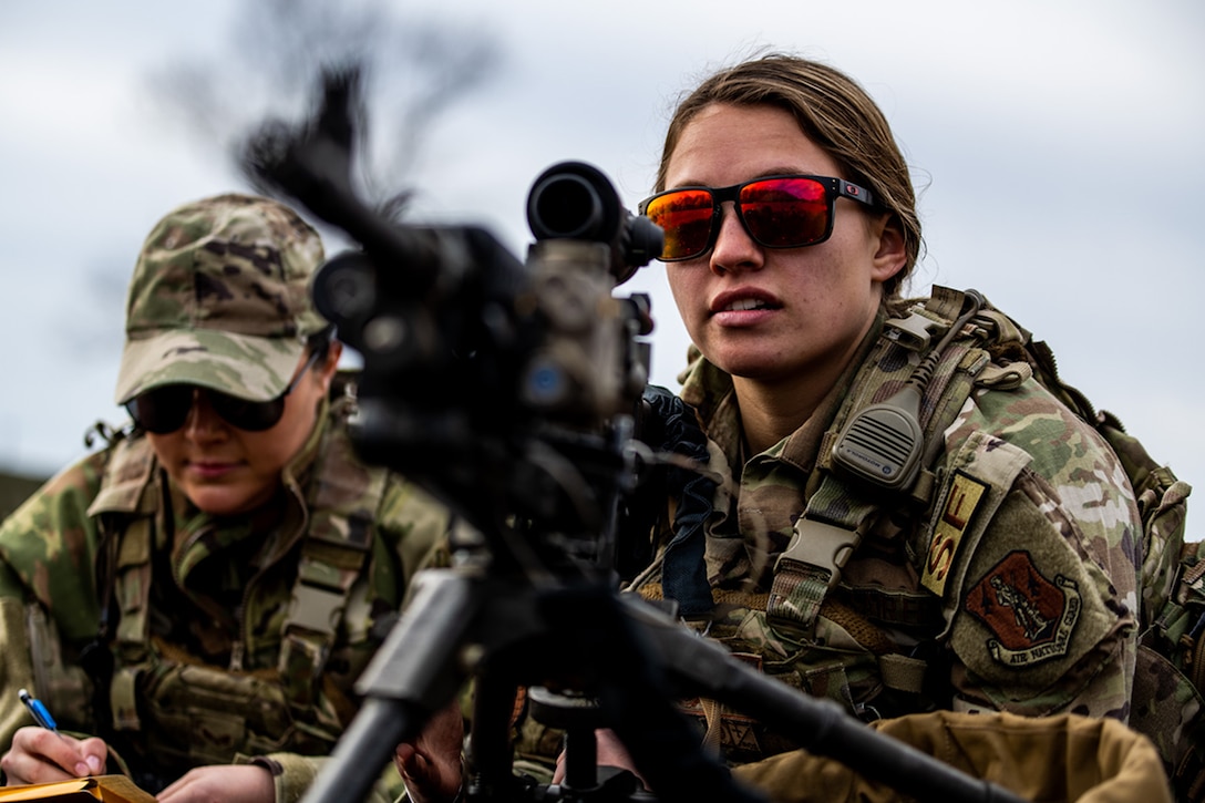 A uniformed service member wearing sunglasses looks through the scope of a weapon as a second takes notes.