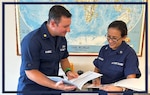 A photo of two Coast Guard members, one officer, and one enlisted, in front of a world map, holding paperwork and in the action of going over the contents contained within the documents.