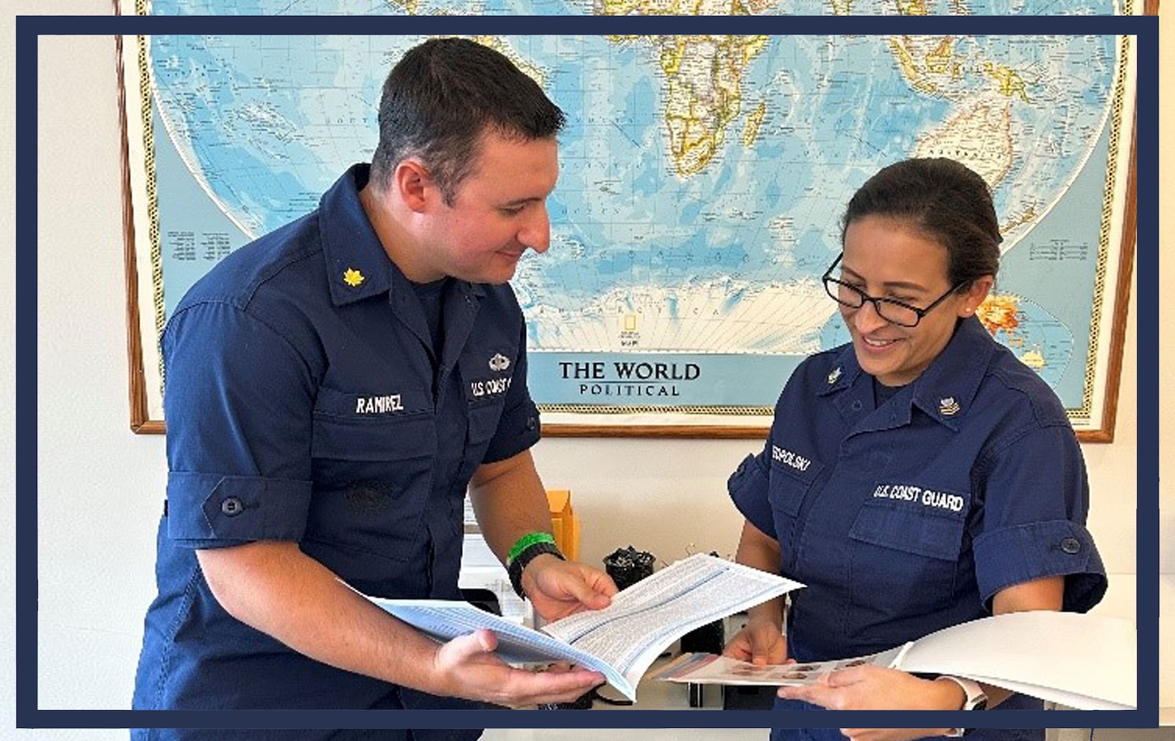 A photo of two Coast Guard members, one officer, and one enlisted, in front of a world map, holding paperwork and in the action of going over the contents contained within the documents.