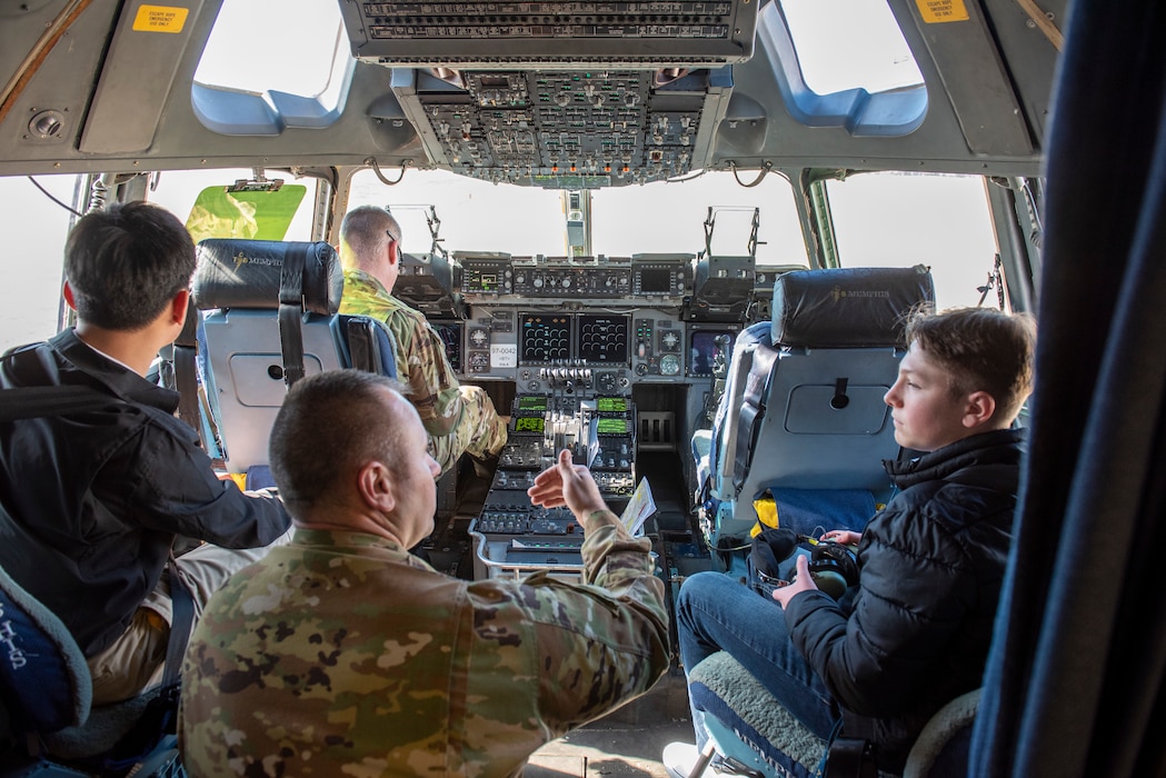 two young men sit in jump seats in a cockpit as an Airmen kneels and talks to them