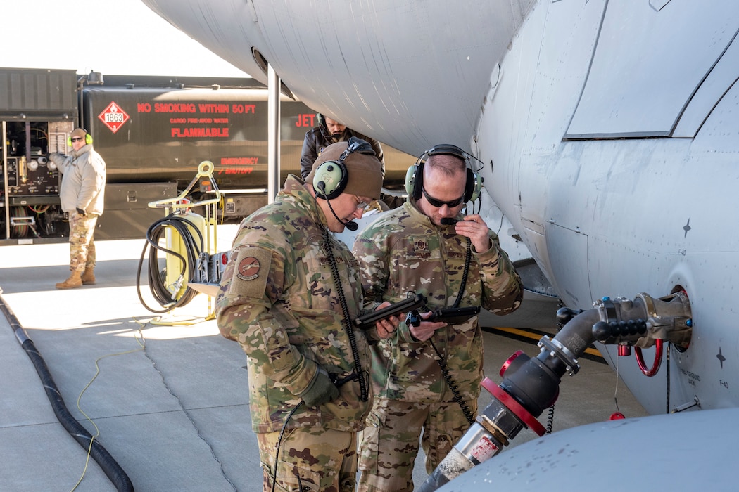 two Airmen look at mobile devices at the side of an aircraft that is receiving fuel