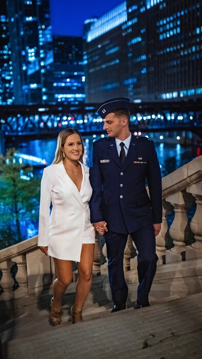 A woman in a white blazer attire is walking up the steps with a man in a formal Air Force uniform.
