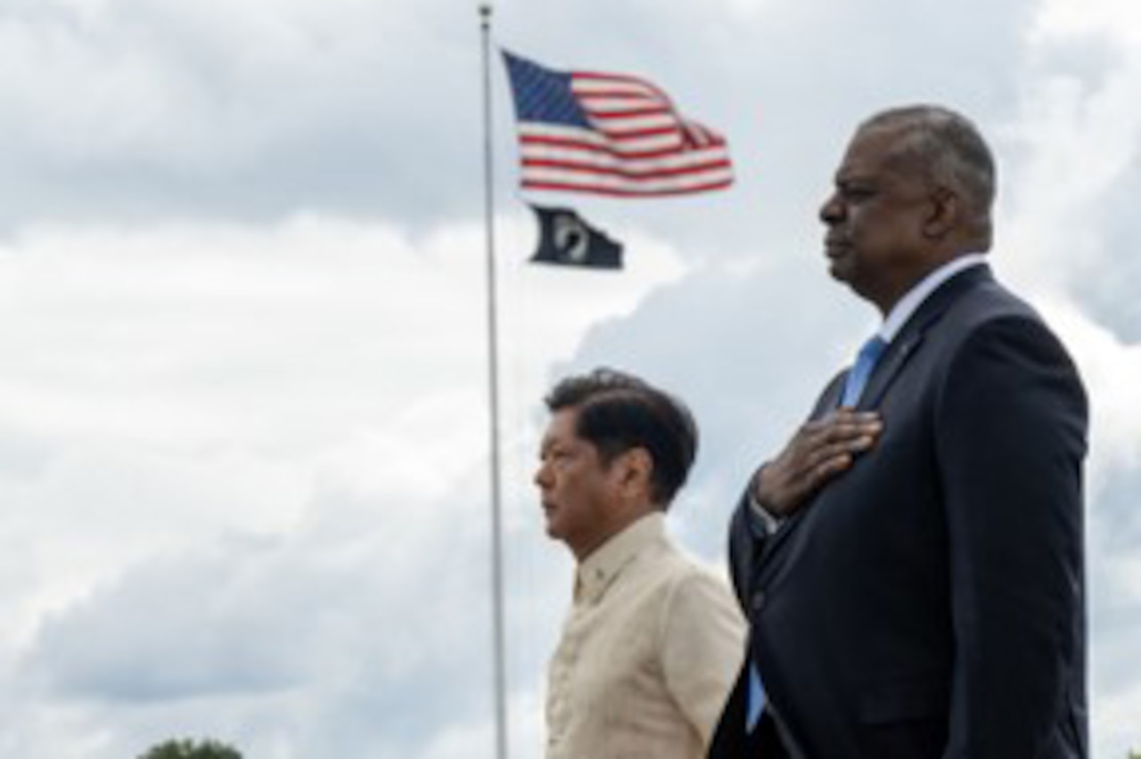 Two people stand at attention. Flags fly in the background.