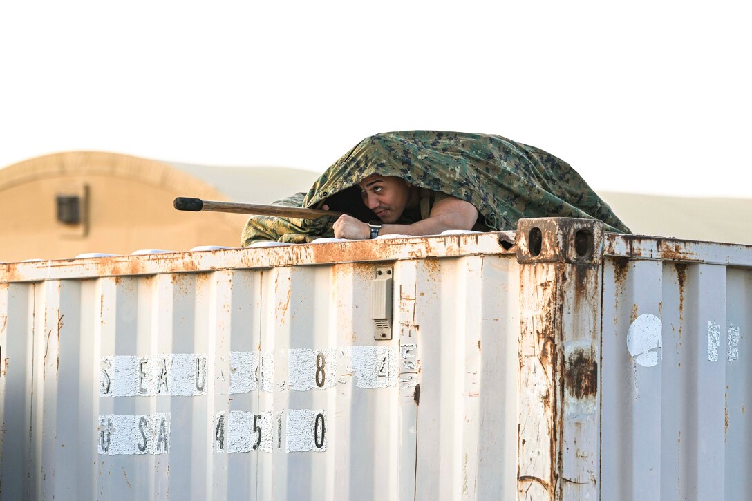 A Marine lays under a camouflage blanket on top of a container while aiming a weapon.