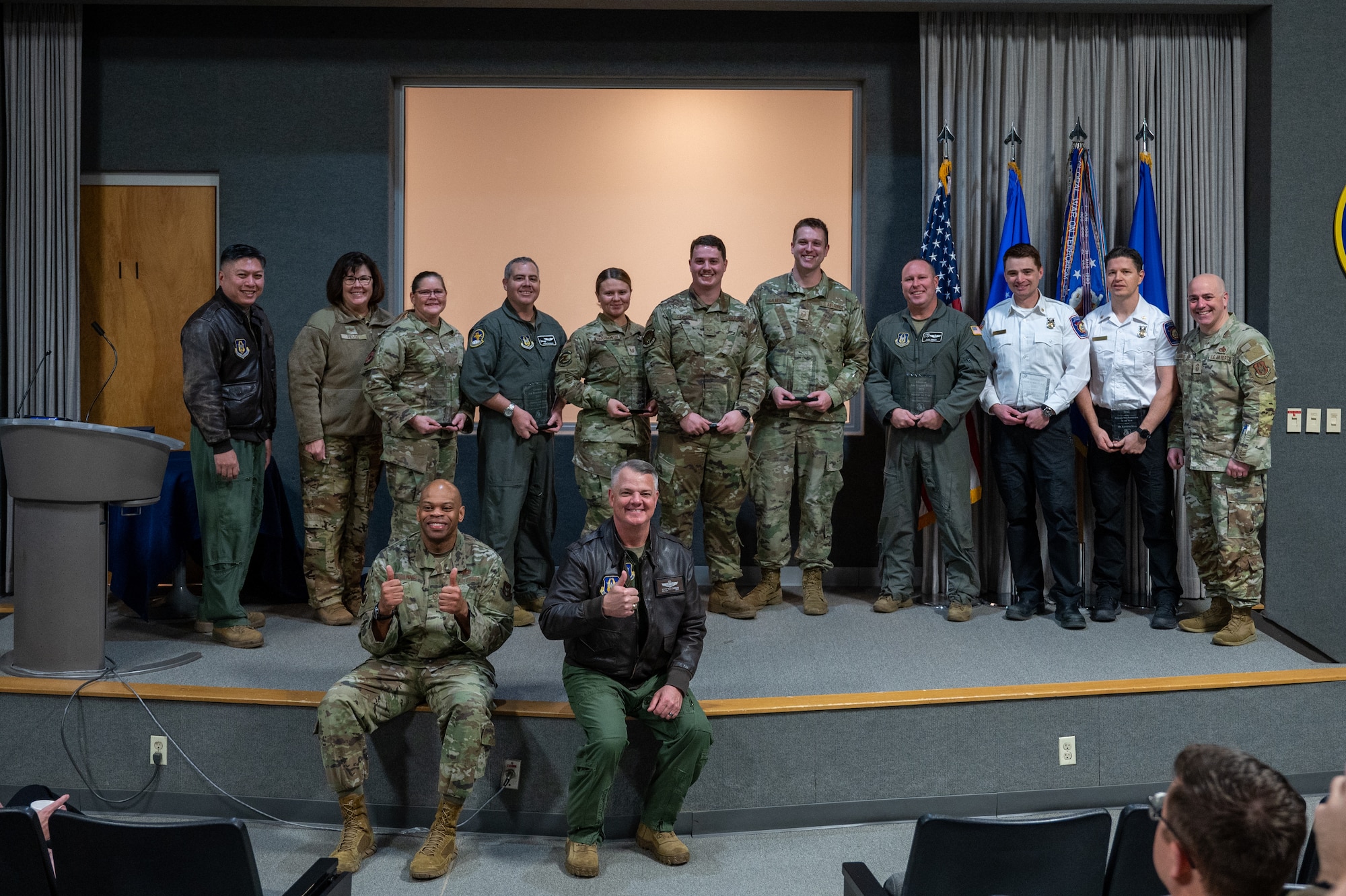 Several men and women stand on a stage, most with awards in their hands. Two men sit at the edge of the stage, smiling for a group photo. All people visible are wearing a variation of a U.S. Air Force uniform.
