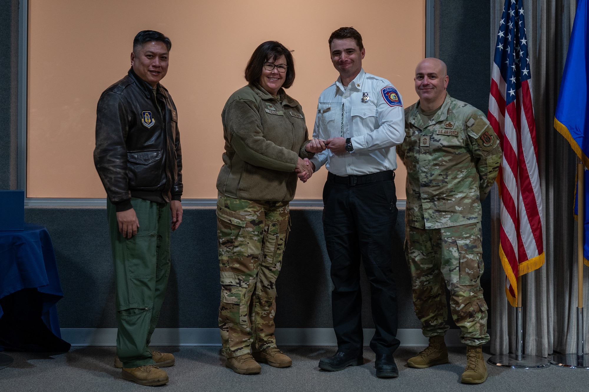A man, with an award in his hand, shakes the hand of a woman. Two other men, on either side of them, pose for a photo with the award. All are dressed in various U.S. Air Force uniforms.