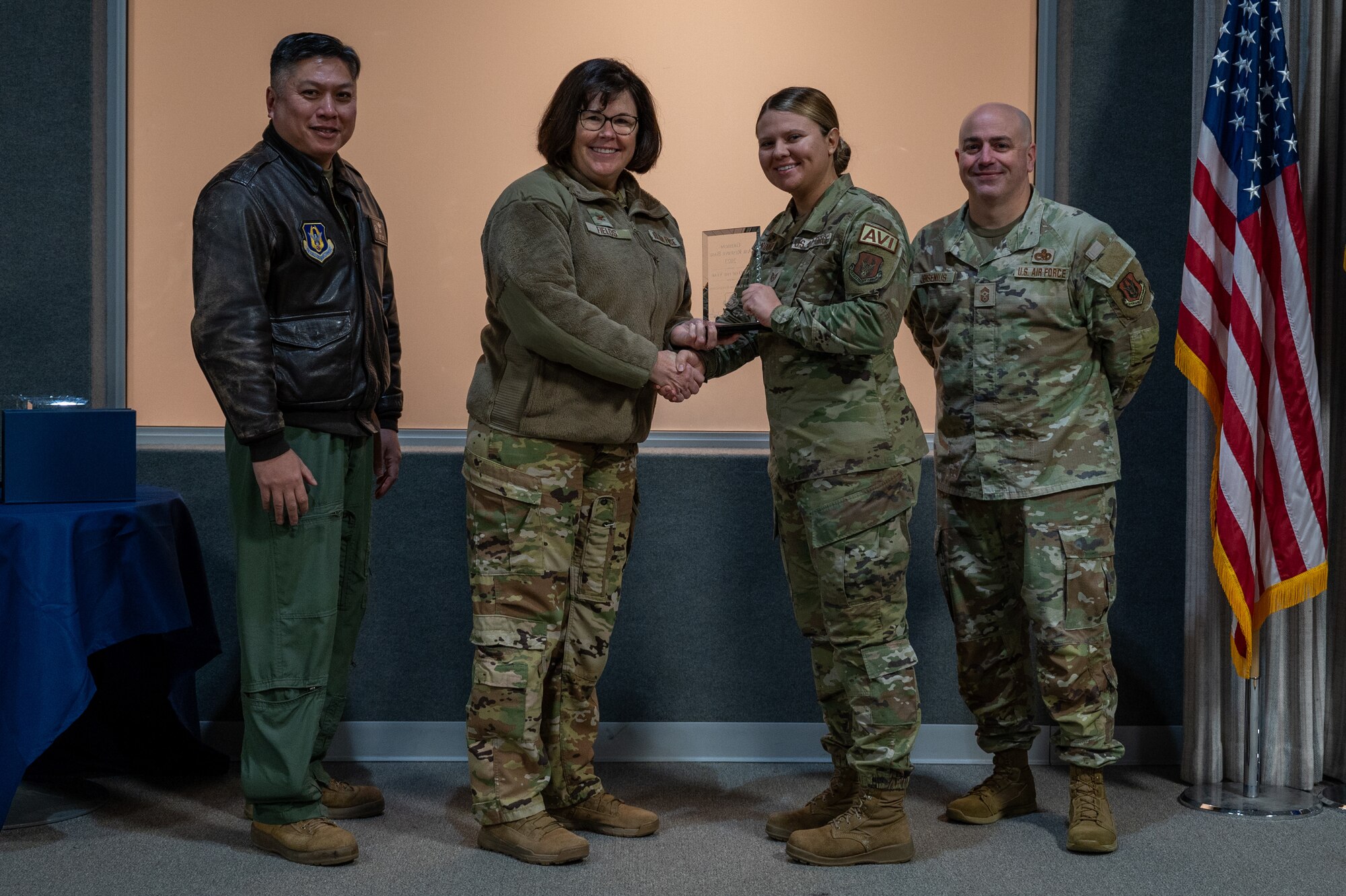 A woman, with an award in her hand, shakes the hand of another woman. Two other men, on either side of them, pose for a photo with the award. All are dressed in various U.S. Air Force uniforms.