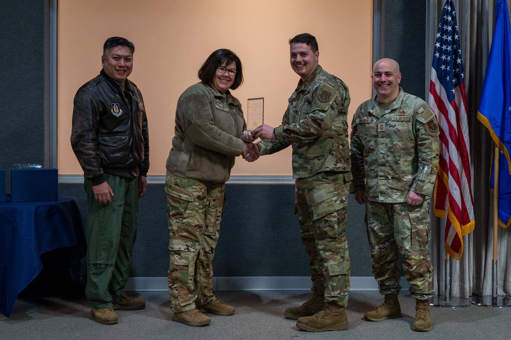 A man, with an award in his hand, shakes the hand of a woman. Two other men, on either side of them, pose for a photo with the award. All are dressed in various U.S. Air Force uniforms.