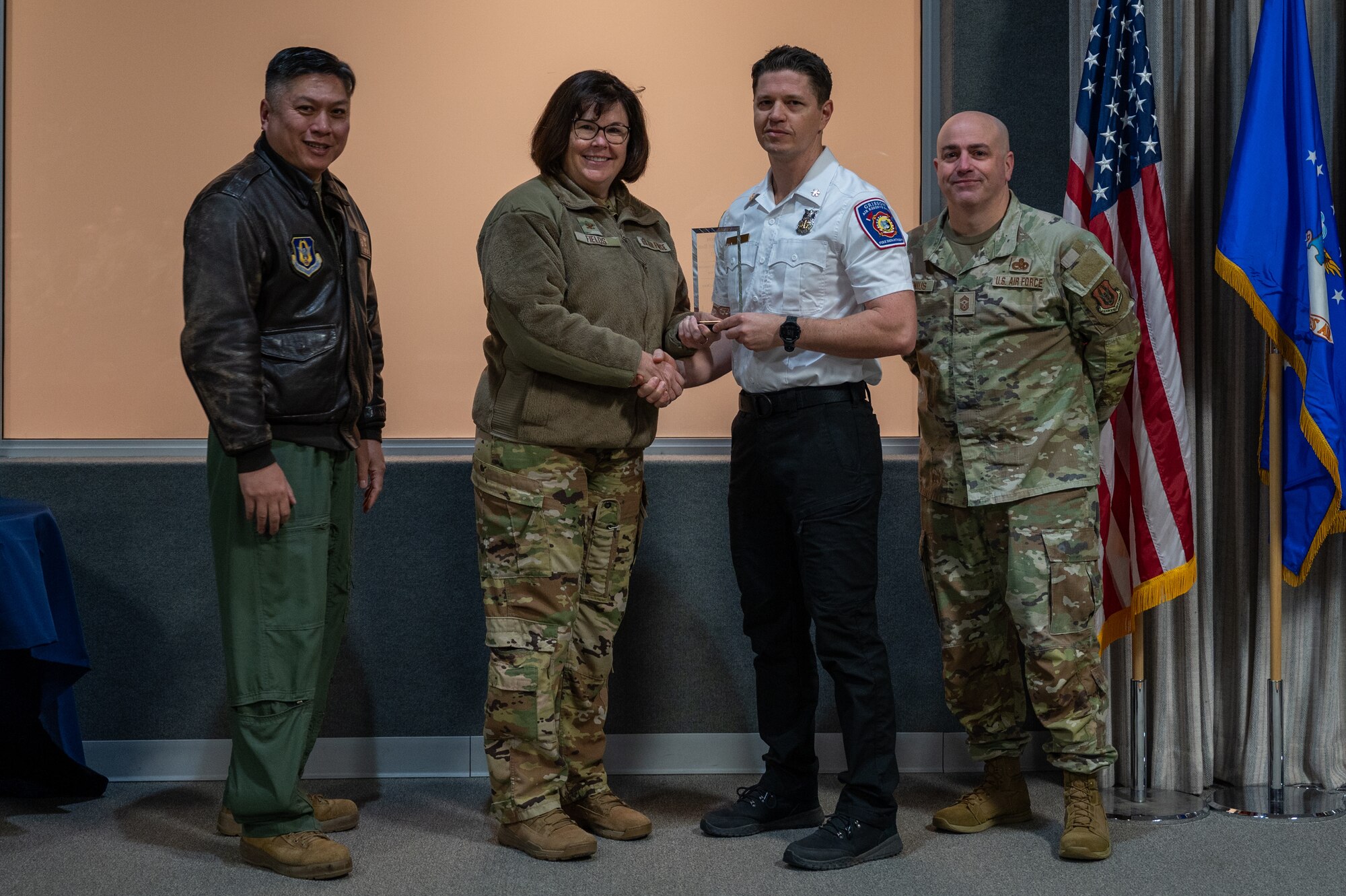 A man, with an award in his hand, shakes the hand of a woman. Two men, on either side of them, pose for a photo with the award. All are dressed in various U.S. Air Force uniforms.