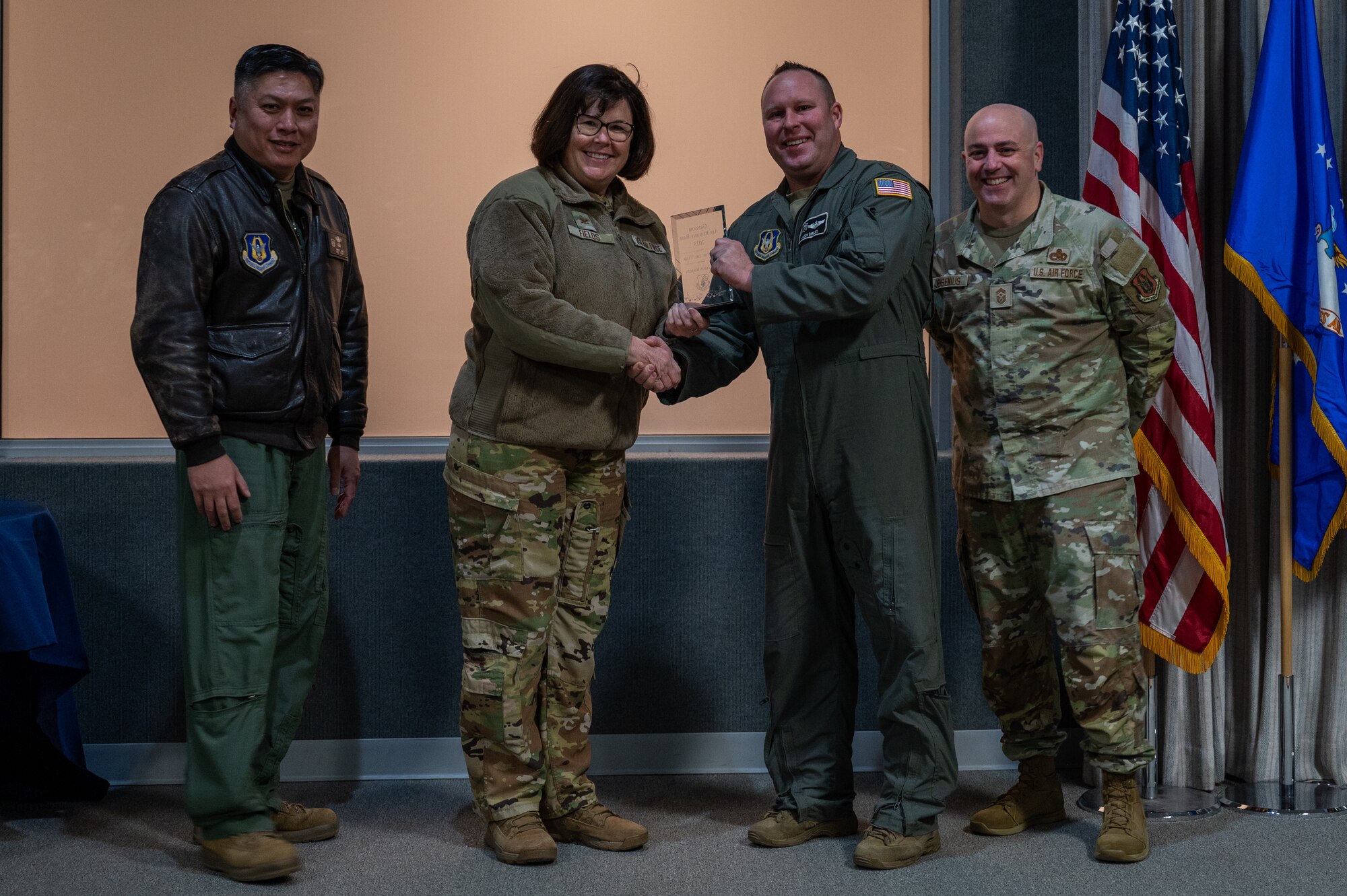 A man, with an award in his hand, shakes the hand of a woman. Two men, on either side of them, pose for a photo with the award. All are dressed in various U.S. Air Force uniforms.
