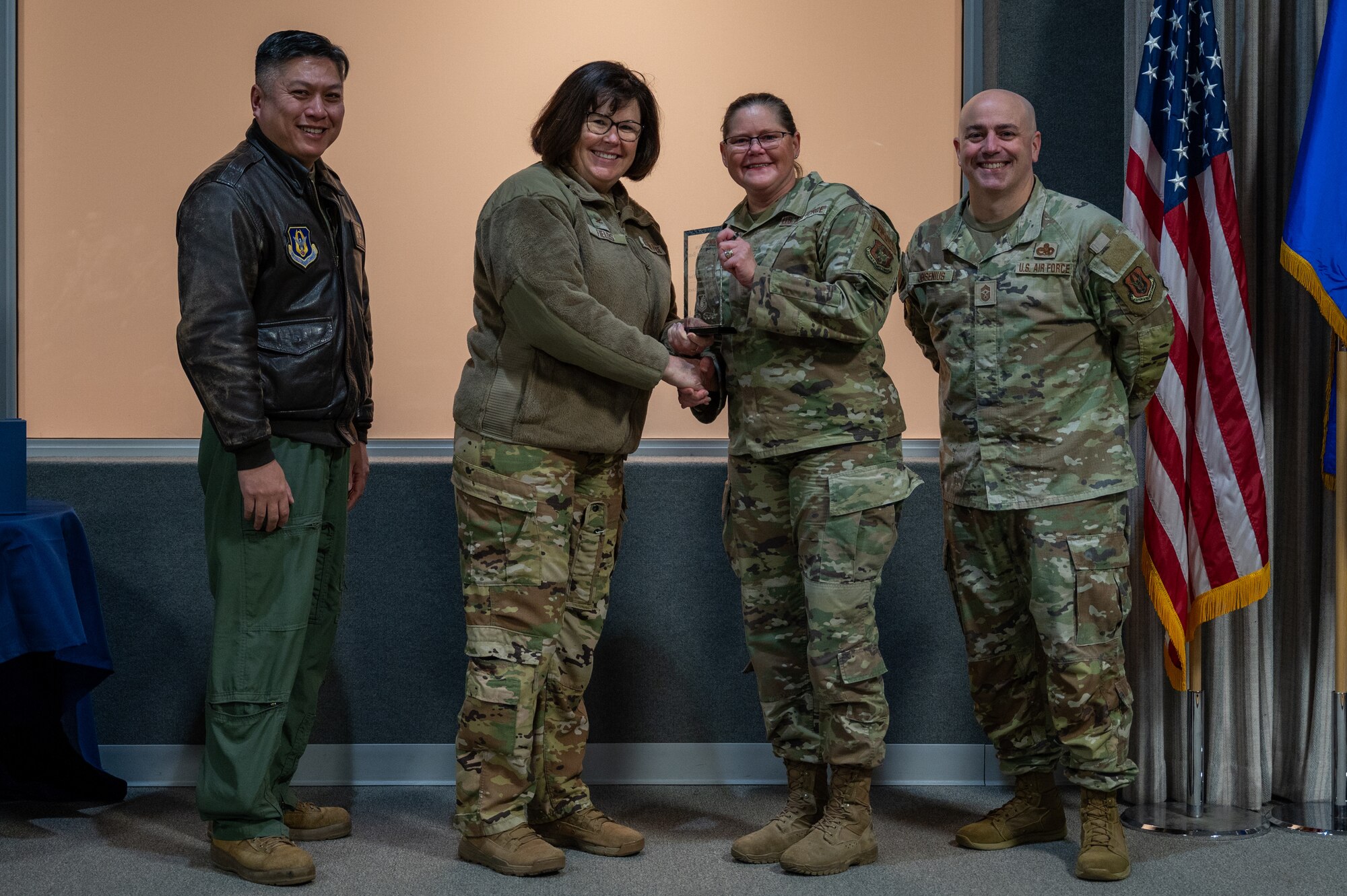 A woman, with an award in her hand, shakes the hand of another woman. Two men, on either side of them, pose for a photo with the award. All are dressed in various U.S. Air Force uniforms.