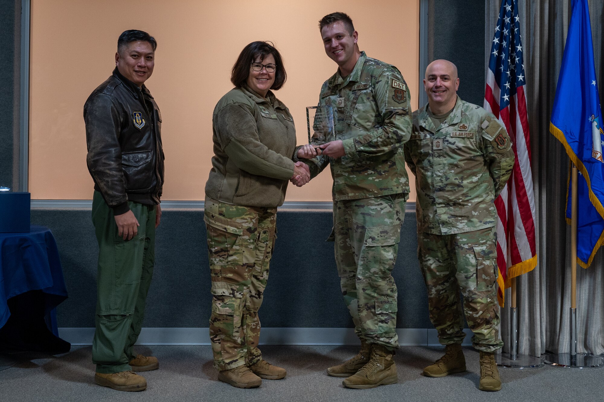A man, with an award in his hand, shakes the hand of a woman. Two other men, on either side of them, pose for a photo with the award. All are dressed in various U.S. Air Force uniforms.