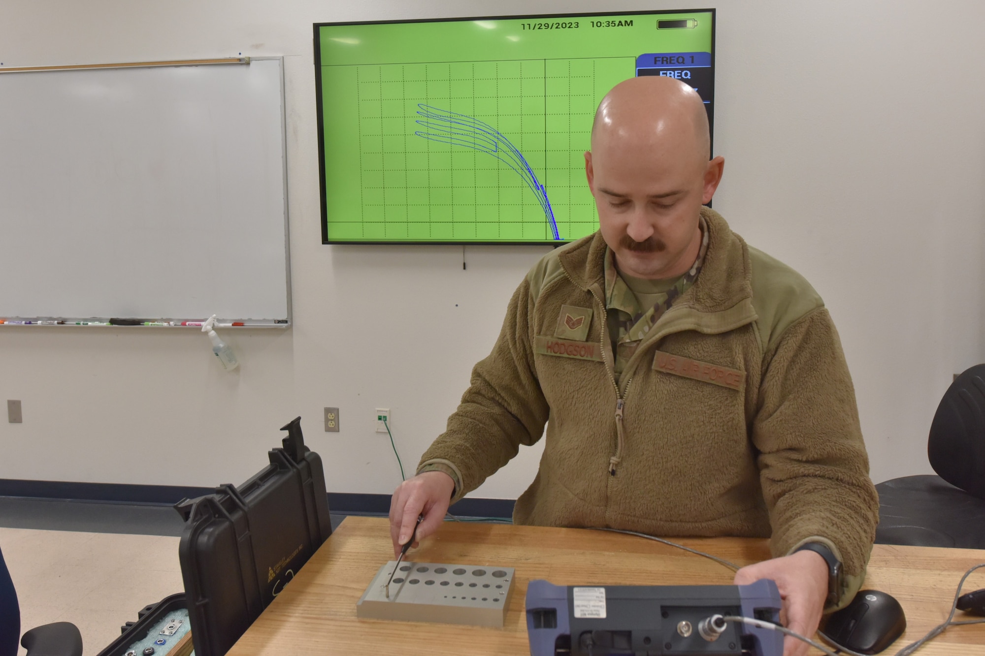An Airman uses an instrument on a piece of metal. The readings are displayed on a monitor behind him.