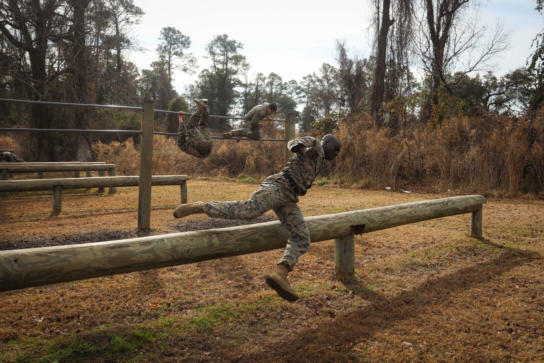 Marines and sailors jump over wooden barriers and climb over metal bars.