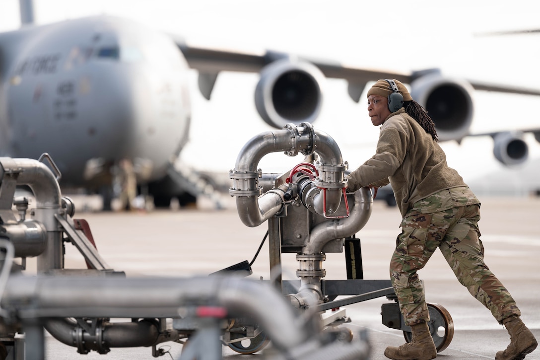 An airman wearing ear protection rolls a system of pipes across the tarmac. A large aircraft is in the background.