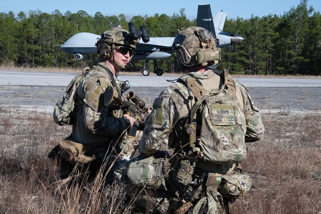 Two Airmen in tactical gear speak to one another while a large military drone sits in the background